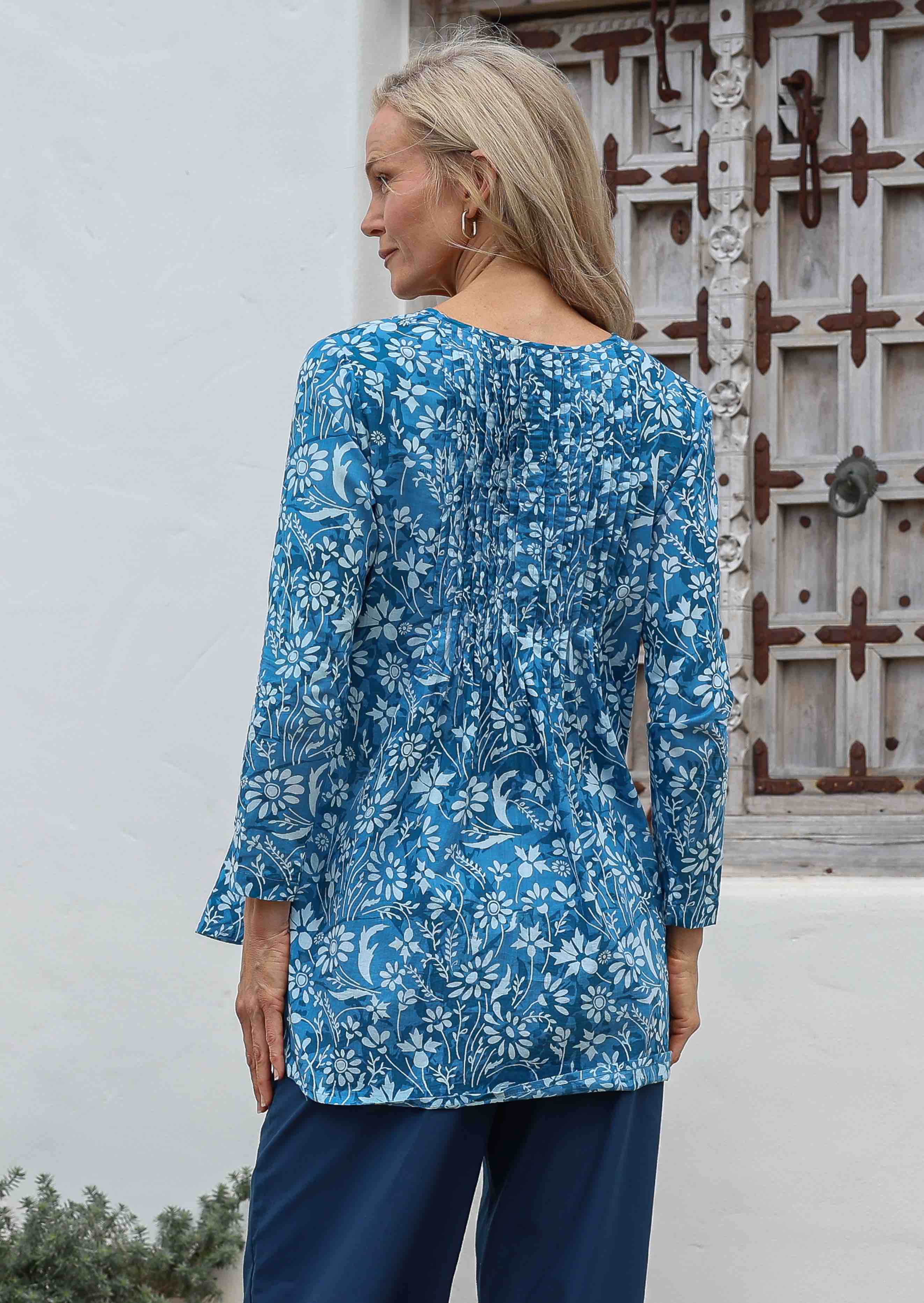 Woman standing in front a wooden window wears 100% cotton pleated front and back detail full sleeve top with V neckline and A line shape.