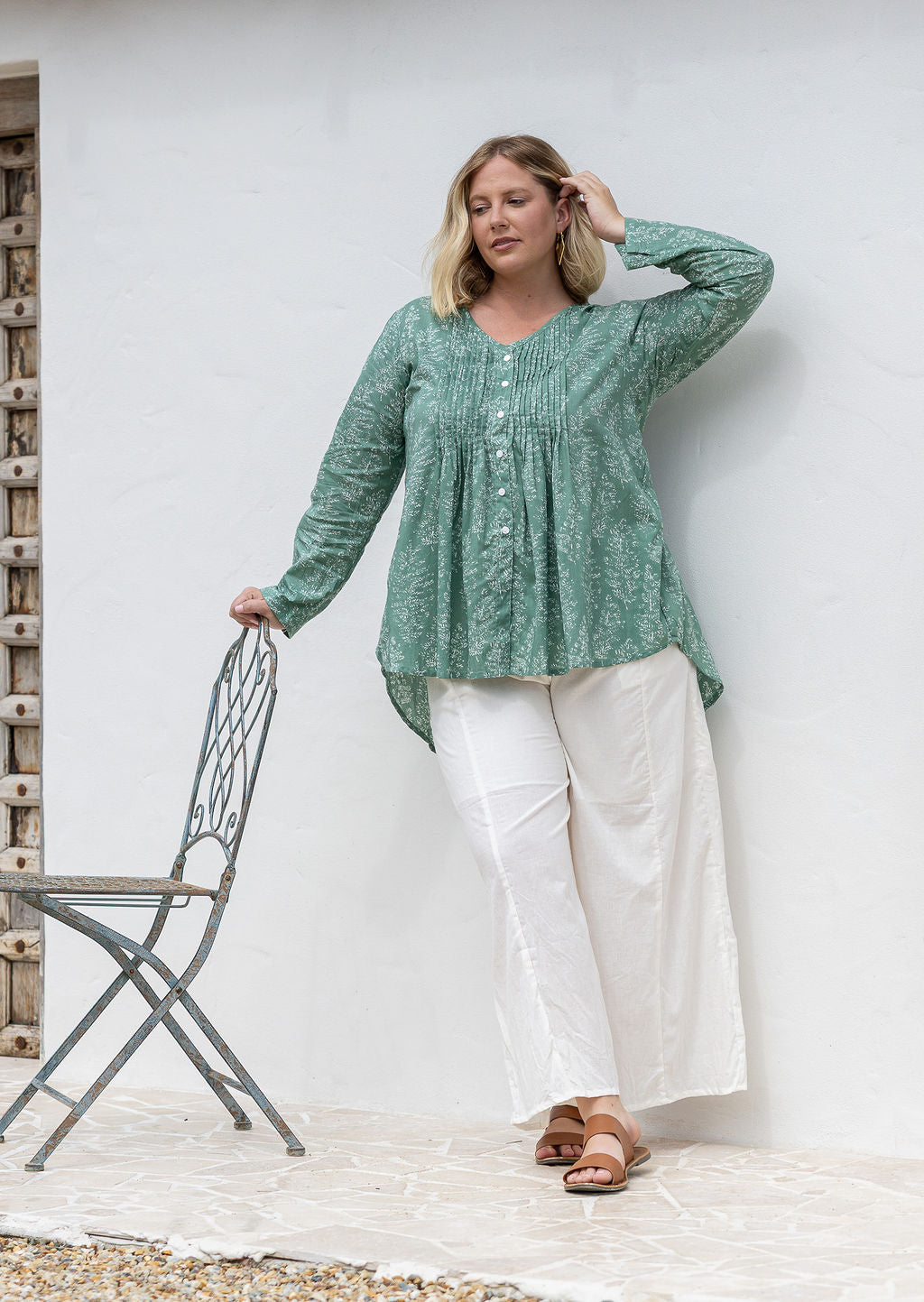 Woman wearing a sage green based floral cotton long sleeve shirt with pin tucks across the back of the bodice, the back of shirt is longer than front