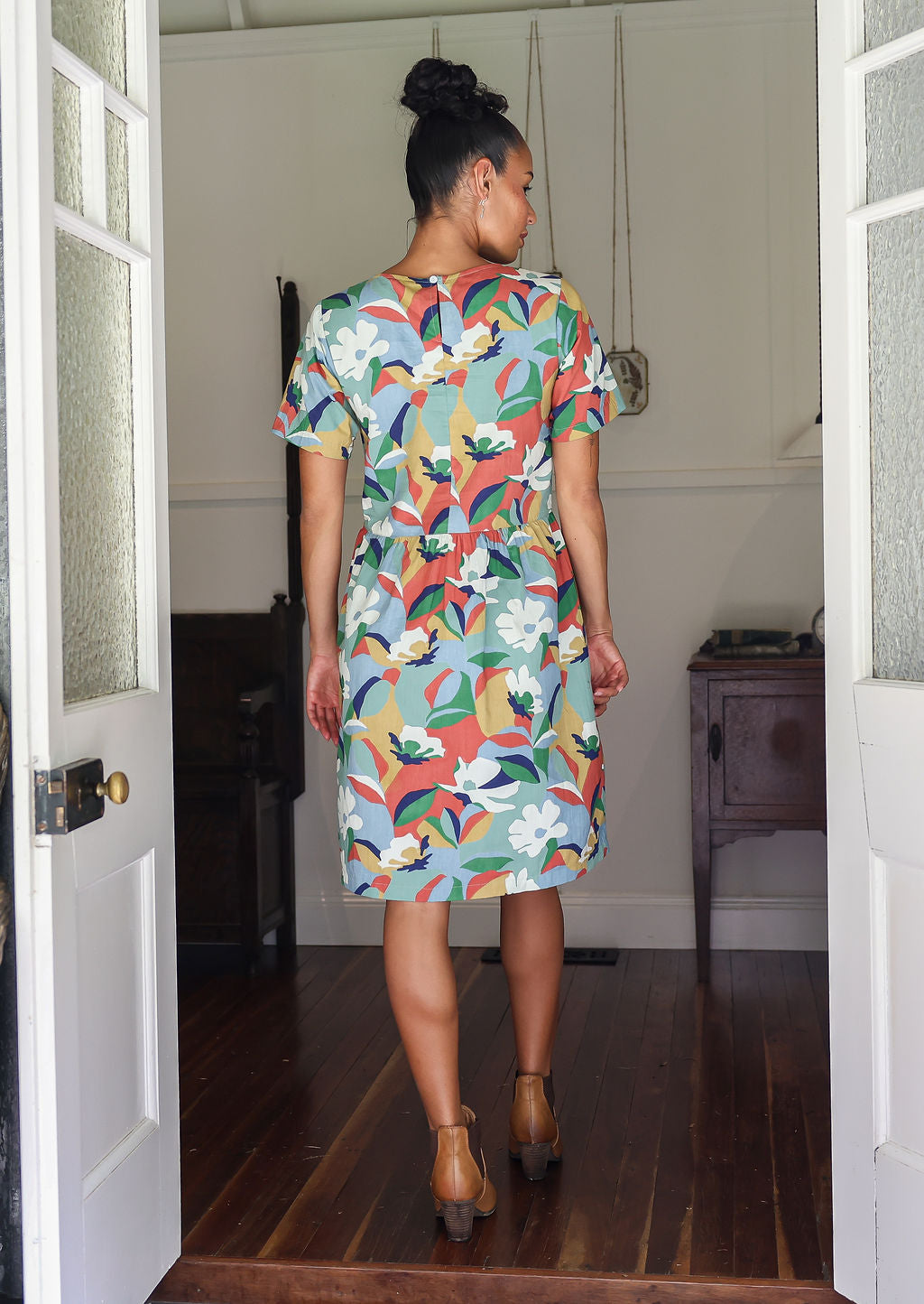 Woman stands backwards wearing a relaxed fit, floral cotton print dress with pockets and empire waistline.