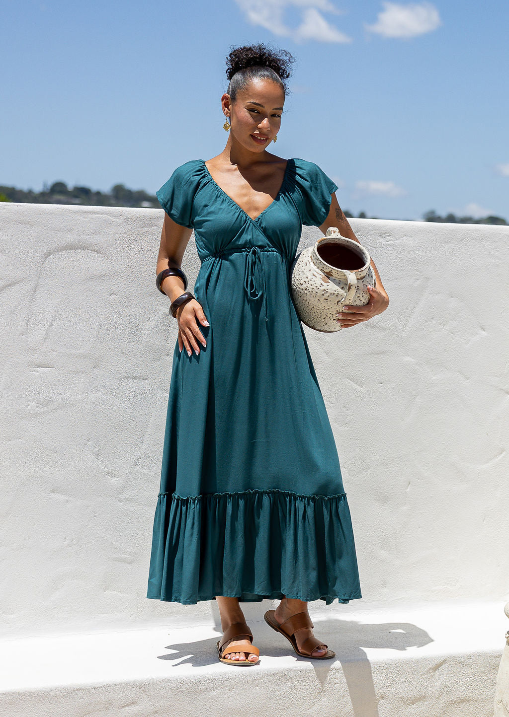 Woman in a teal dress maxi dress holding a ceramic pot against a white wall with blue sky.