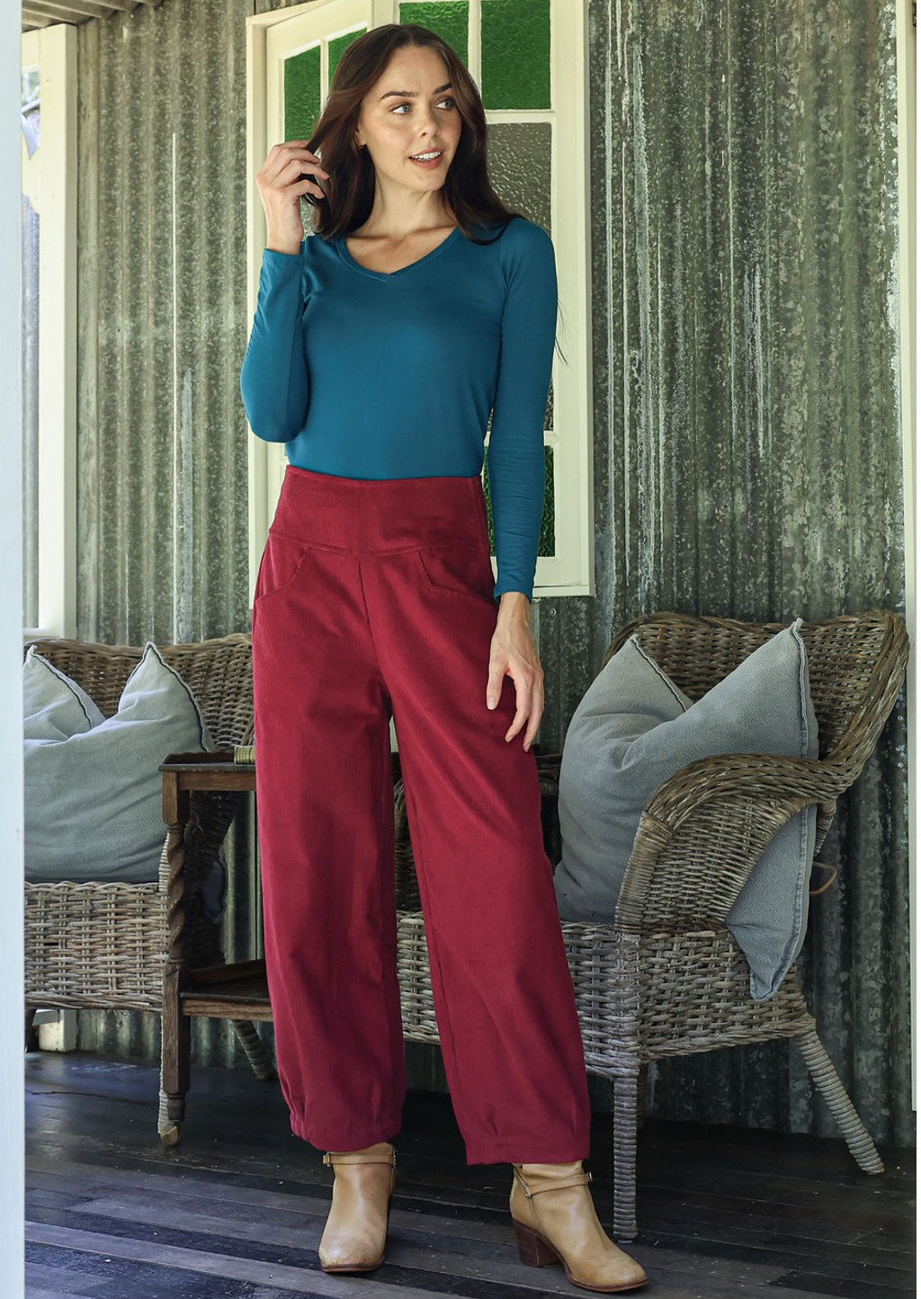 Woman in teal top and red pants standing in a room with wicker furniture and green walls.
