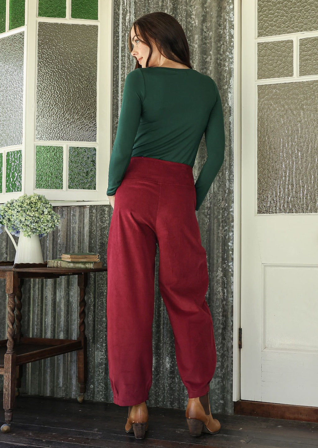 Woman stands backwards wearing a green top and red pants standing in a room with textured walls and a small table with flowers.
