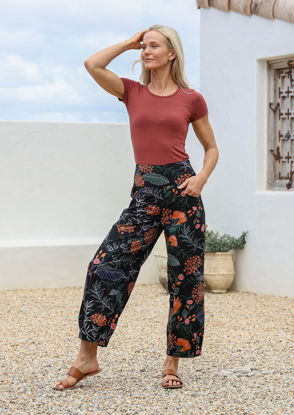 Woman posing in 100% cotton wide-leg black botanical printed pants with wide flat waistband, side zipper and small pleats at the ankles.