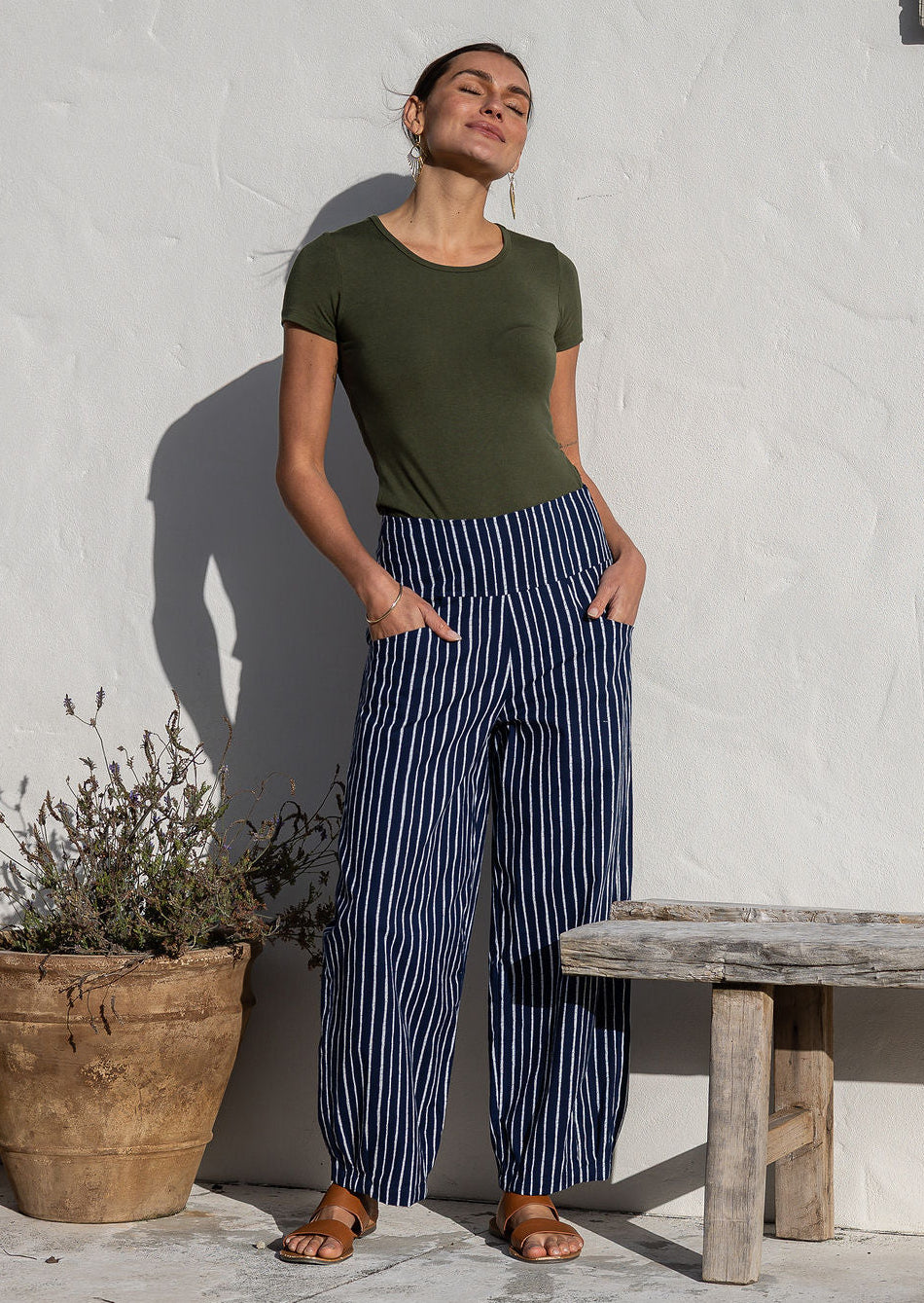 Woman posing in 100% cotton wide-leg navy based white strioed printed pants with wide flat waistband, side zipper and small pleats at the ankles.