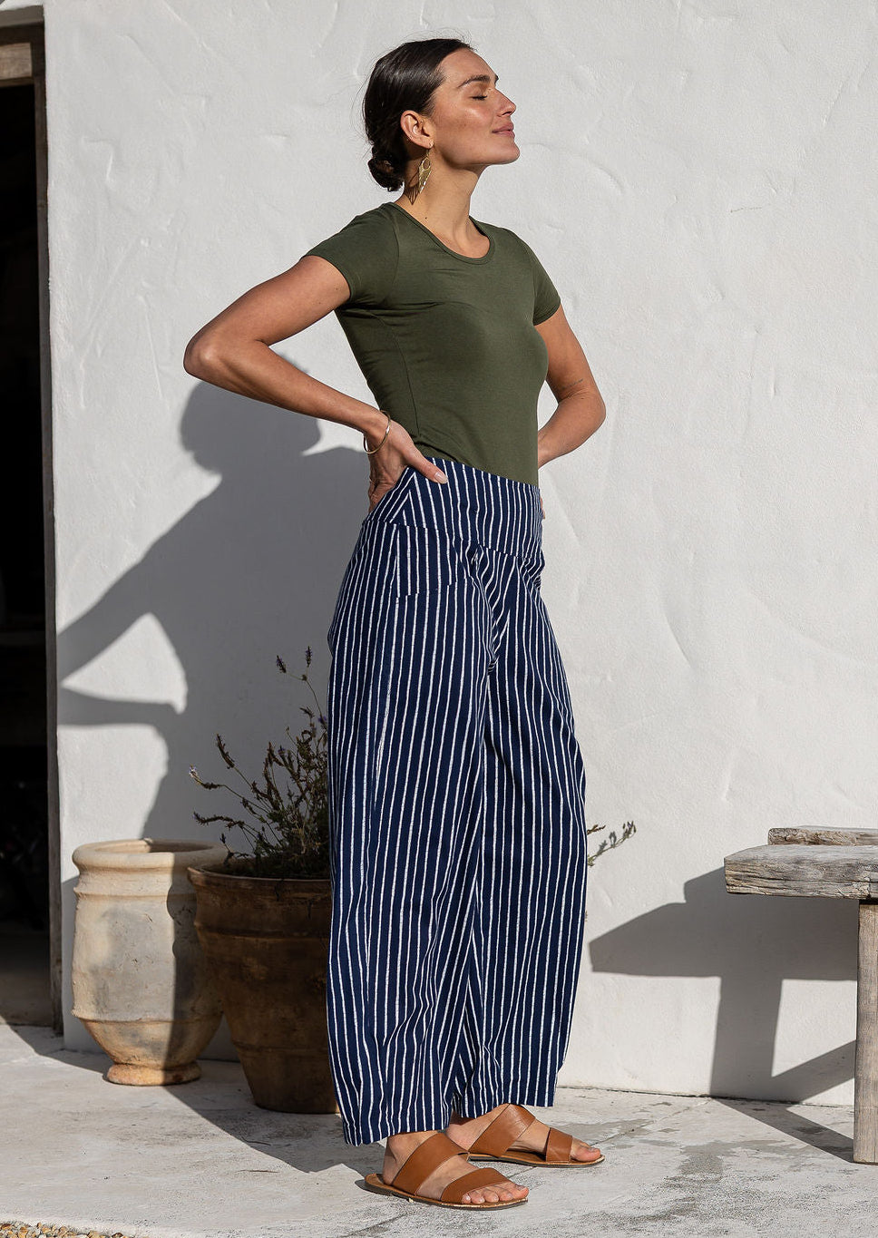 Model standing sideways wears 100% cotton wide leg pants with side zipper, pockets and small pleats at ankles.