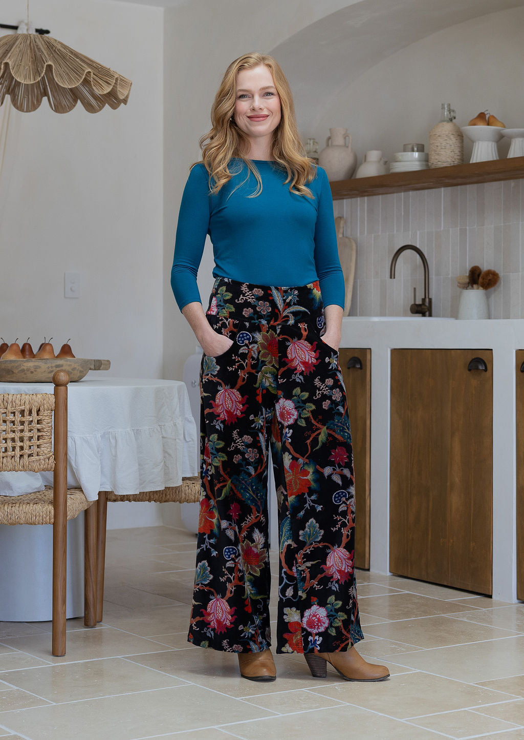 Woman wearing a black based multi-colour floral velvet wide leg pants with both hands in pockets and a side zip