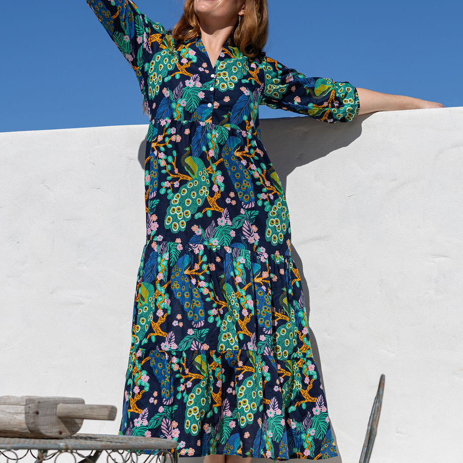 Woman wearing 3/4 sleeved maxi dress with a colourful floral and peacock print in shades of green, blue, yellow, and pink, posing outdoors against a white wall and blue sky.