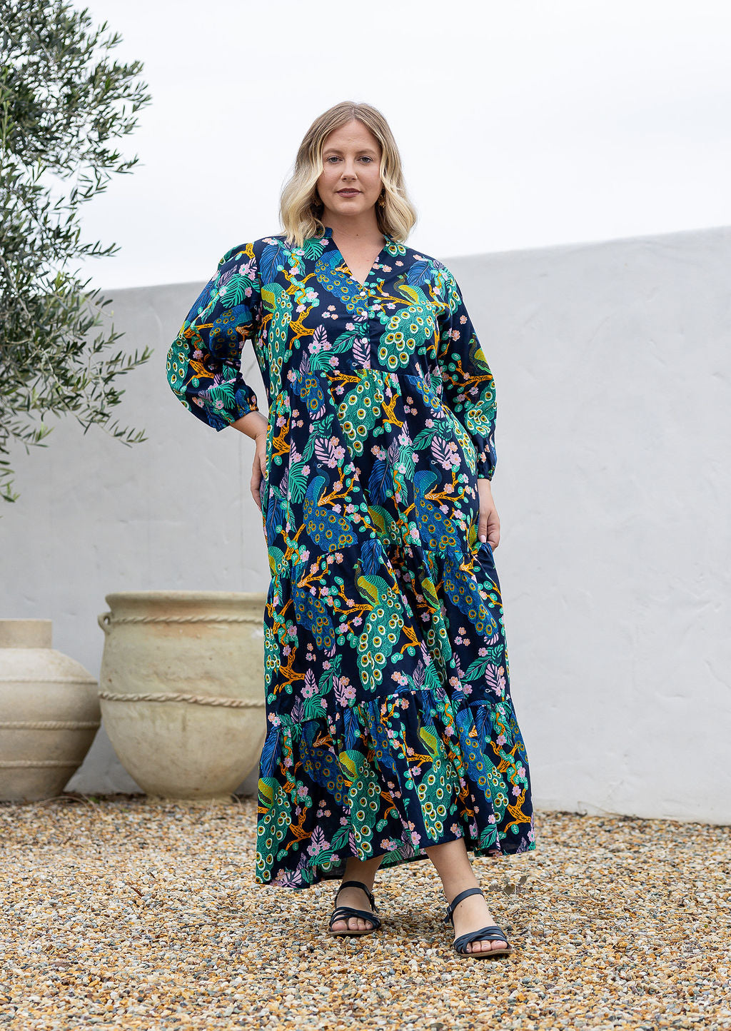 A woman wearing a 3/4 sleeved, buttoned bodice cotton maxi dress with a multicolour floral and peacock print.