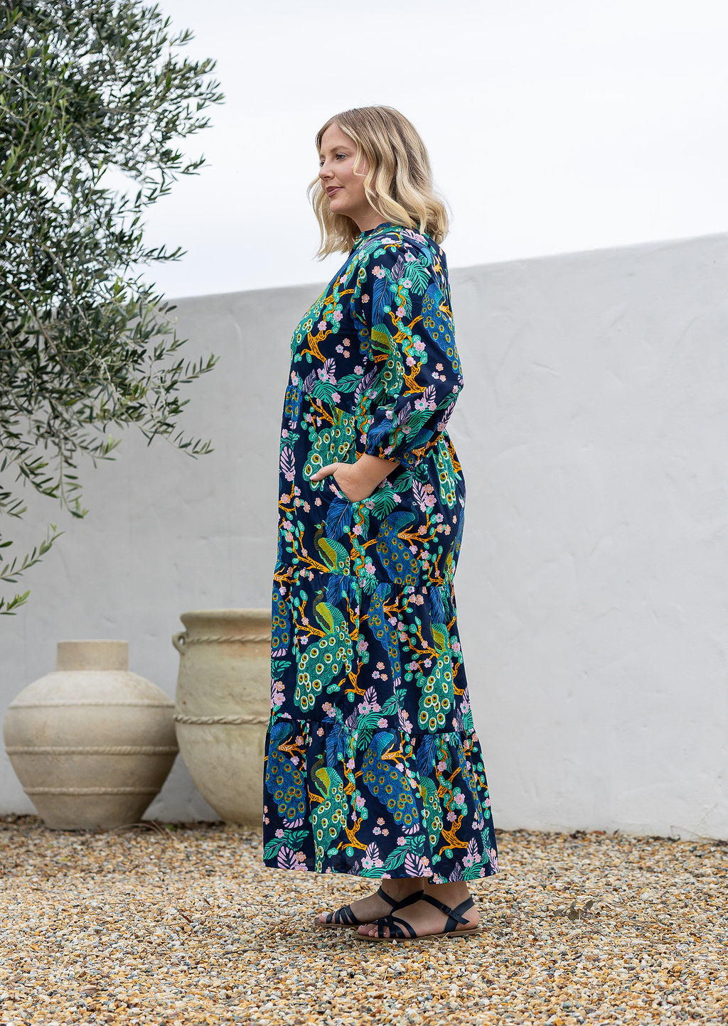 A woman stands sideways wearing a 3/4 sleeved cotton maxi dress with a colourful floral and peacock print in teal, green, yellow, and pink.