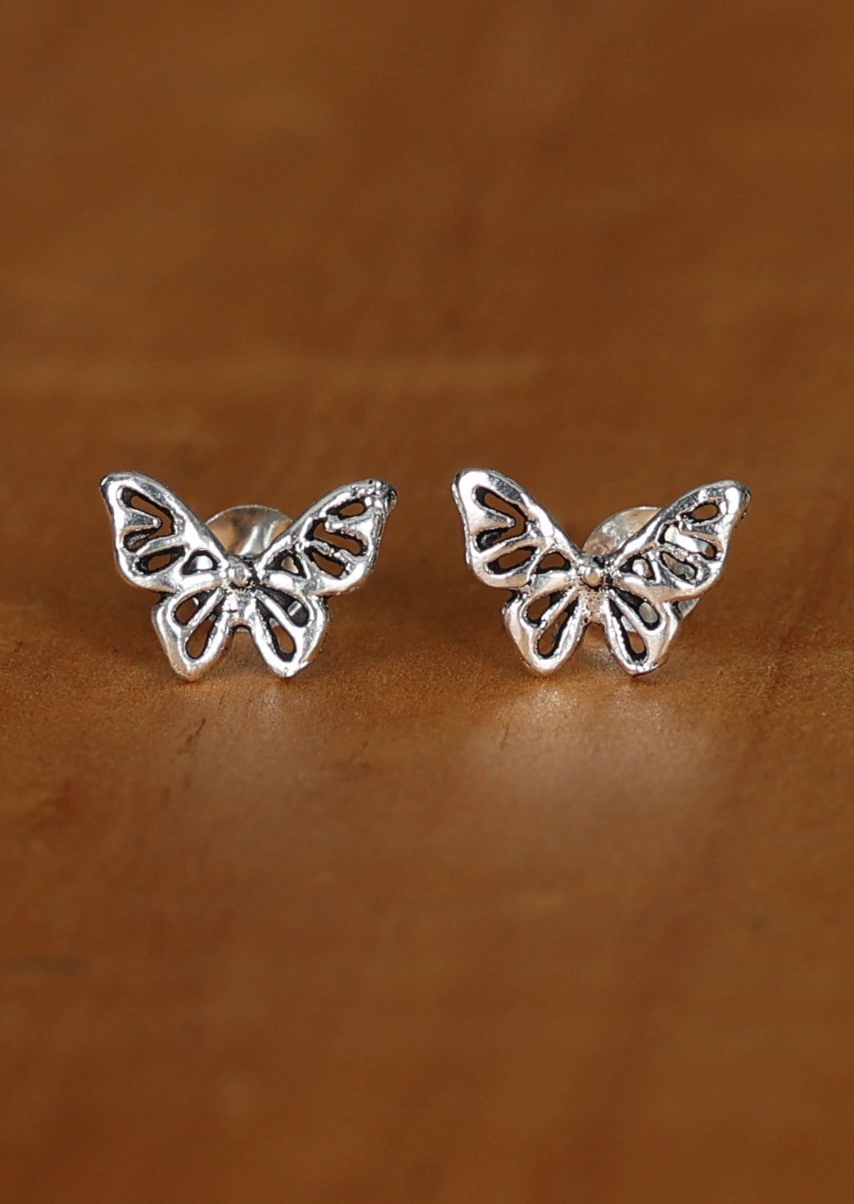 A pair of silver Ethereal butterfly stud earrings with intricate cutout details, displayed against a wooden background.