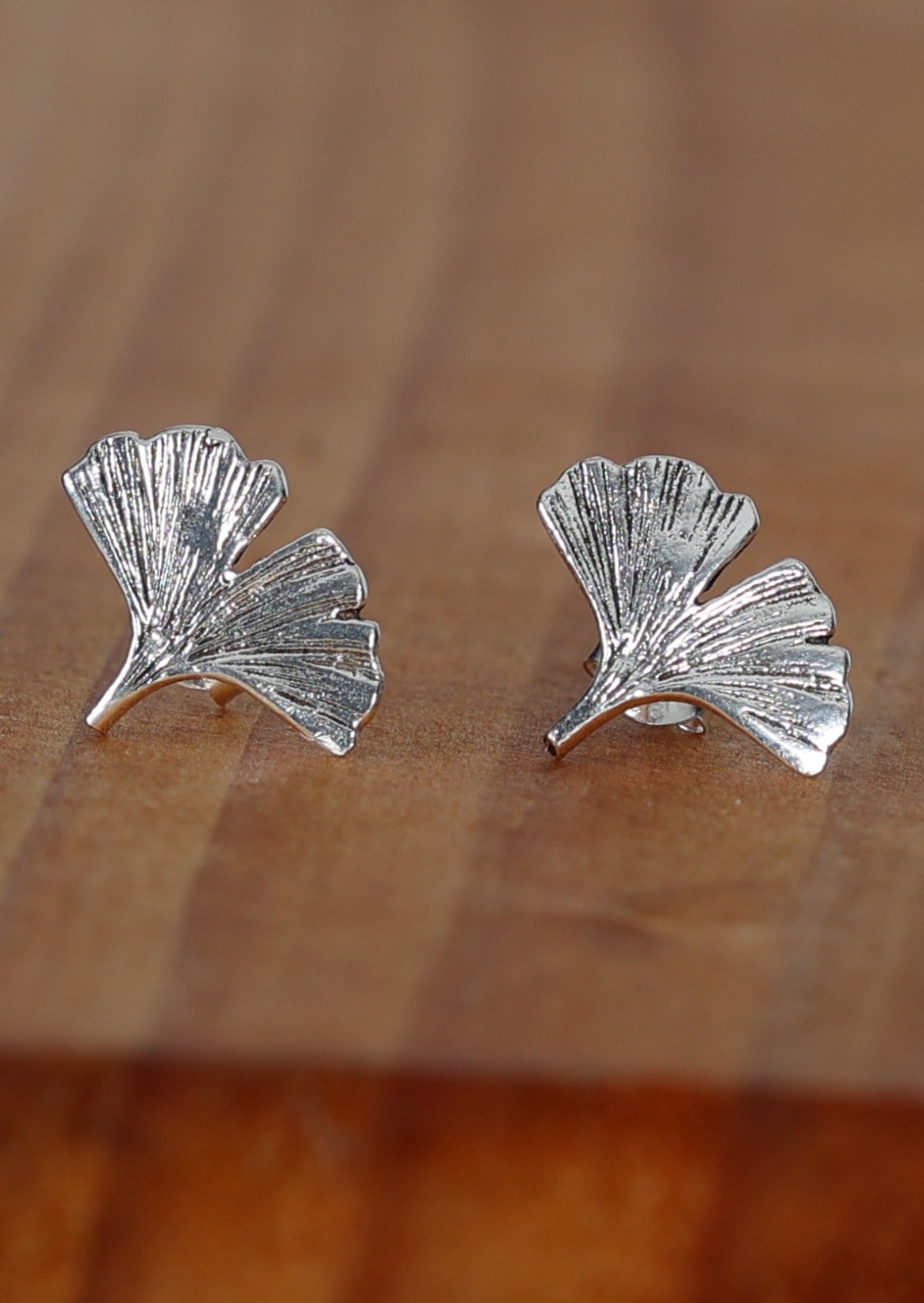 A pair of sterling silver ginkgo leaf stud earrings displayed on a wooden surface.