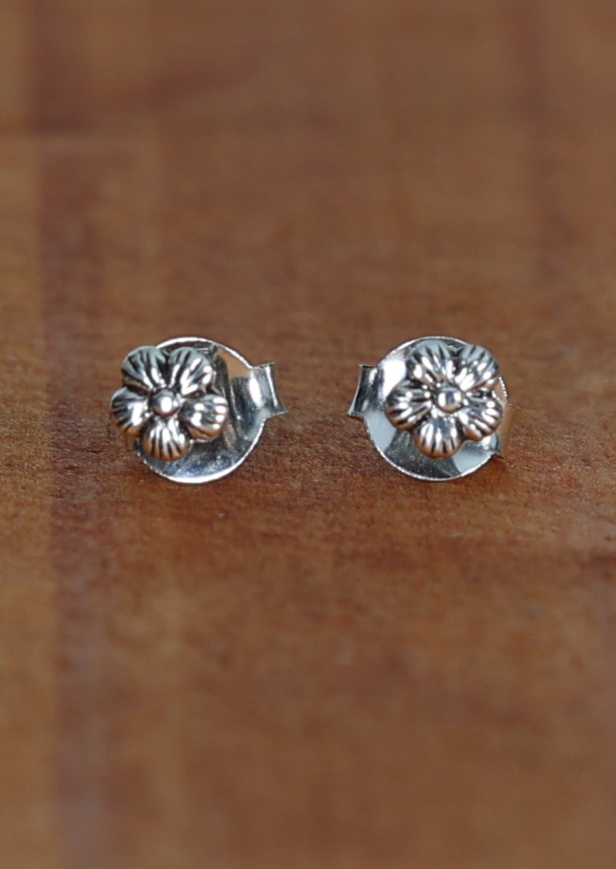 A pair of dainty flower silver studs with delicate bloom design, displayed on a wooden surface