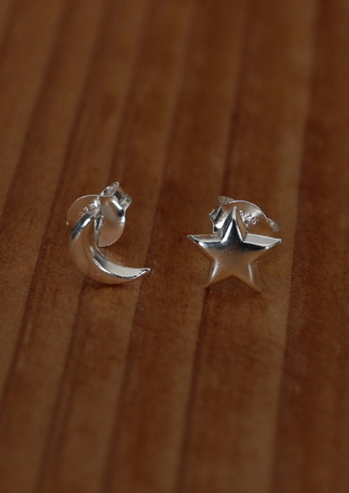 A pair of silver stud earrings, one designed with a crescent moon and the other with a star, placed on a wooden surface.