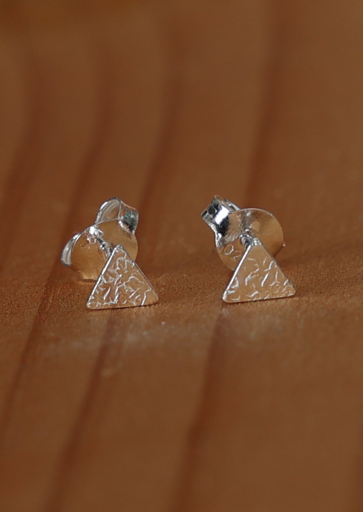 A pair of silver triangle stud earrings with a textured design, displayed against a wooden background.