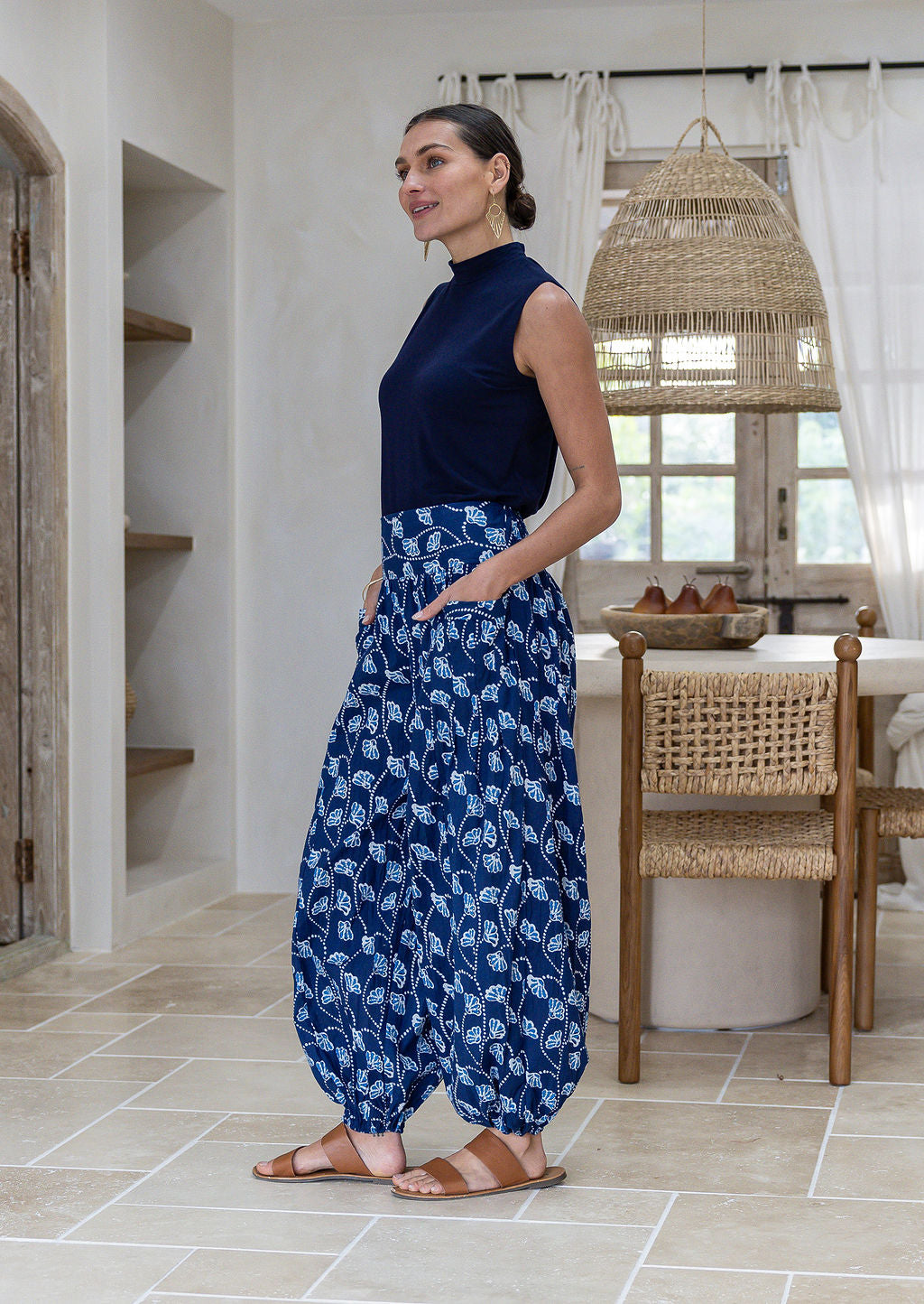 woman standing sideways wearing a 100% cotton floral blue harem pants with flat wide waistband in front with both hand in pockets