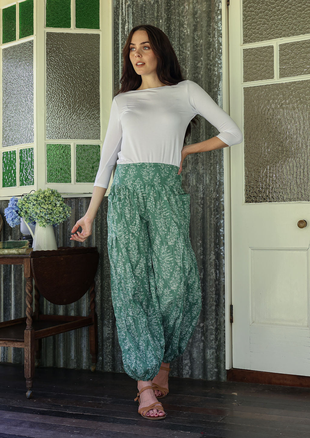 Woman wearing a white top and sage green floral pants with wide elastic waist and ankles standing in a rustic room