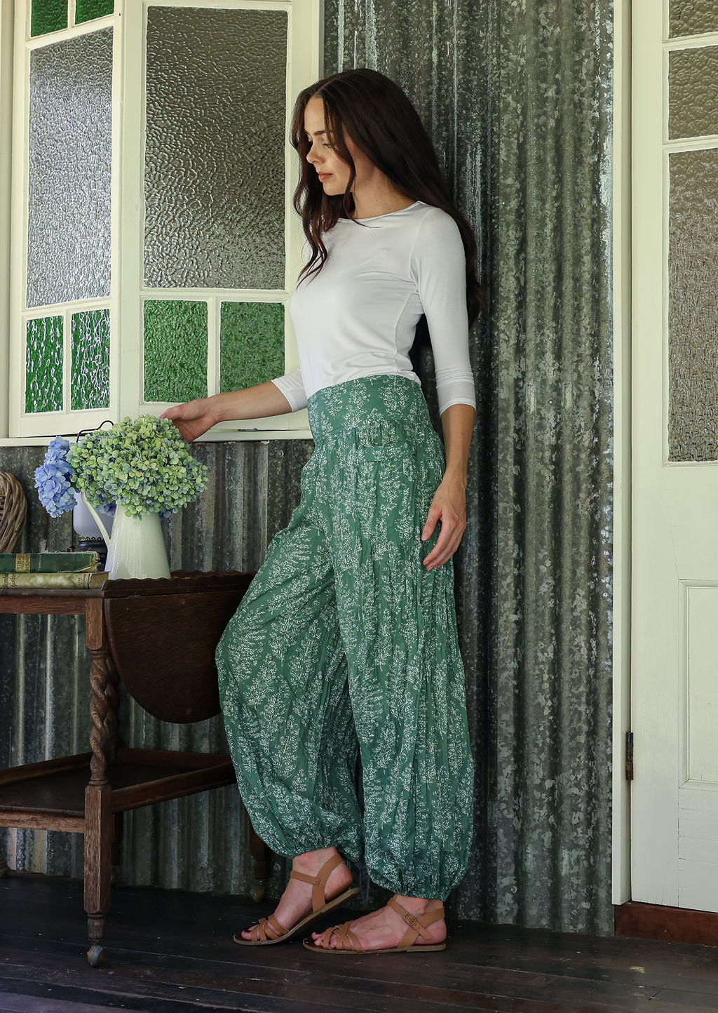 Woman in a white top and sage green based floral pants standing in a room with a window and curtain.