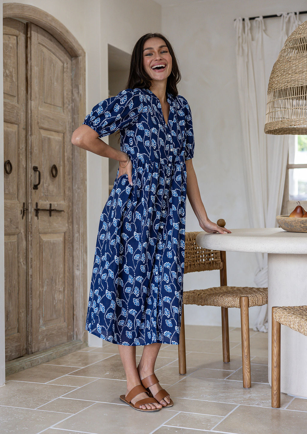 A woman wearing a navy blue based floral midi dress stands in a kitchen setting. The dress features puff sleeves and side pockets.