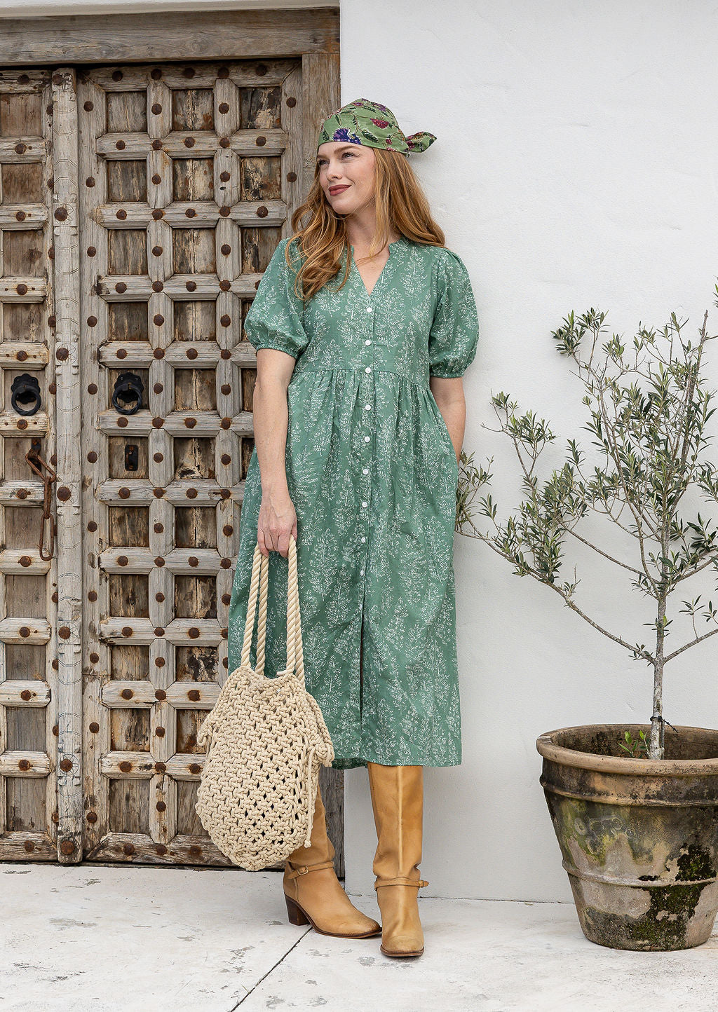 Woman in a sage green based floral print dress standing next to a wooden door and potted plant