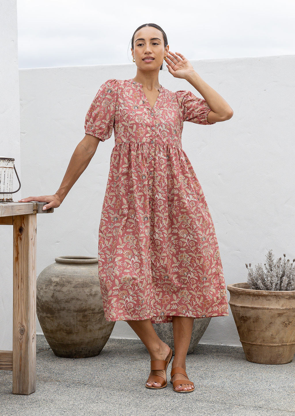 Woman wearing a pink floral cotton dress standing in an outdoor setting with plants and a table.