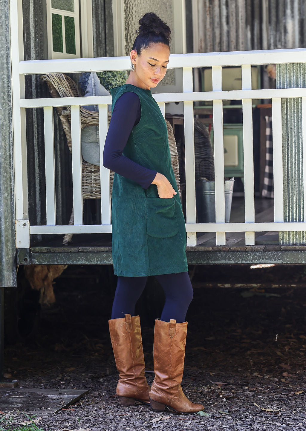Woman stands sideways wearing a green dress and brown boots standing in front of a white railing.