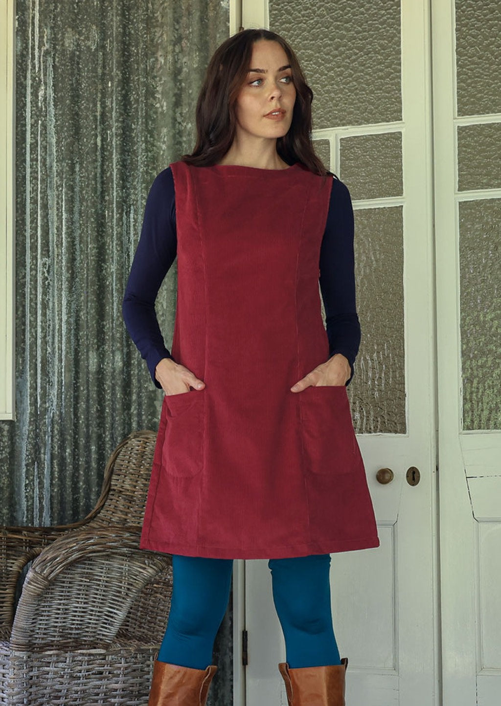 Woman in a red cotton corduroy sleeveless tunic and blue leggings standing in a room with a white door
