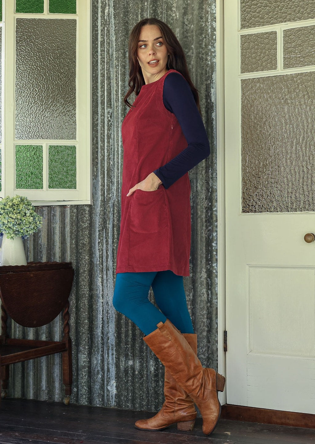 Woman stands sideways wearing a red dress and blue tights standing in a room with patterned walls and a door.