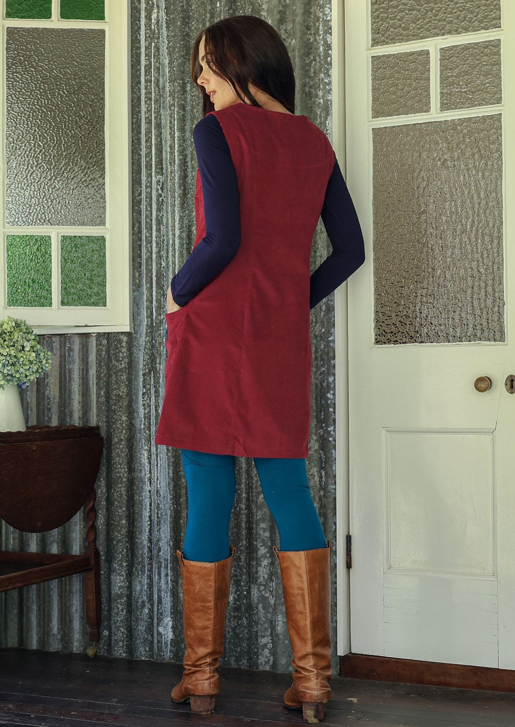 Woman stands backwards wearing a red dress and blue leggings standing in a room with patterned curtains and a white door.