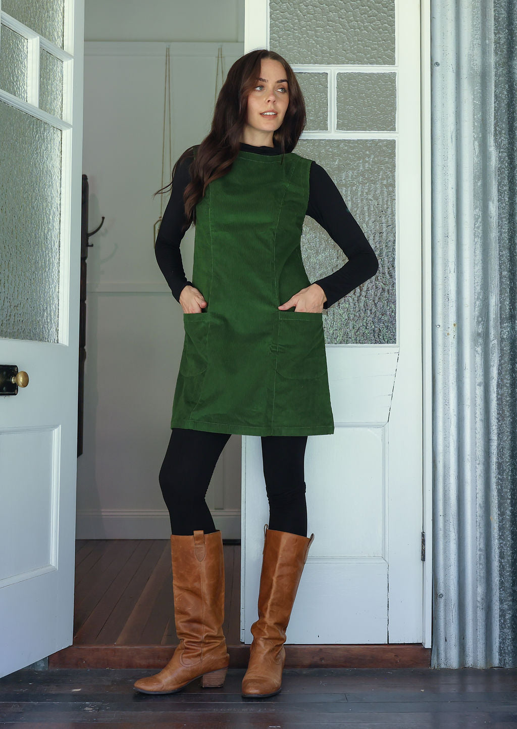 Woman wearing a green cotton corduroy sleeveless tunic layered over a black top and leggings and tan boots, with hands in front pockets
