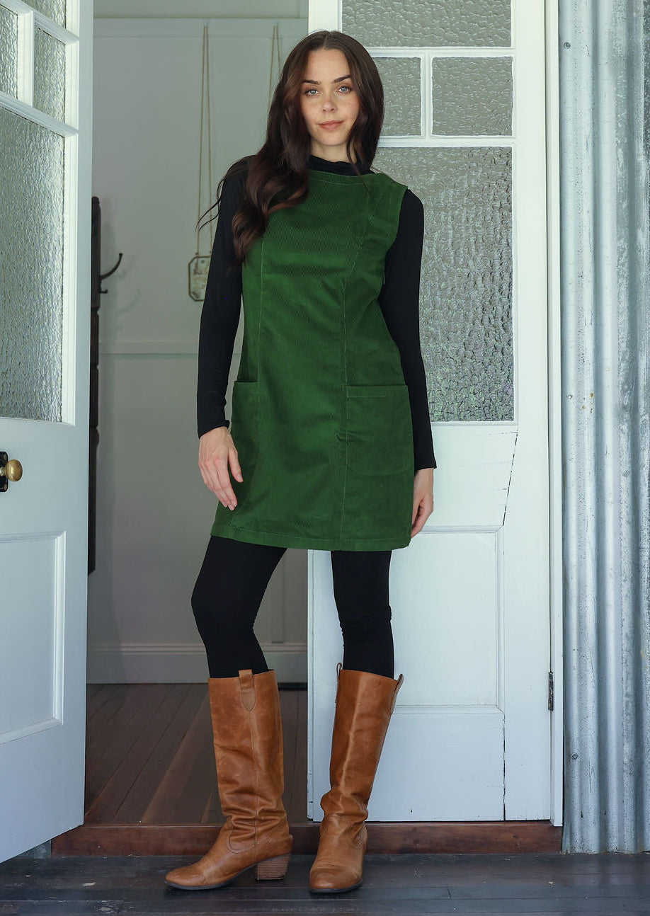Woman wearing a green cotton corduroy sleeveless tunic layered over a black top and leggings and tan boots