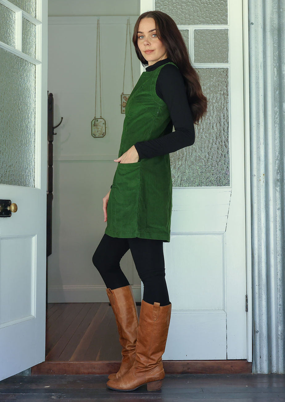 Woman standing side on in a green cotton corduroy sleeveless tunic layered over a black top and leggings and tan boots
