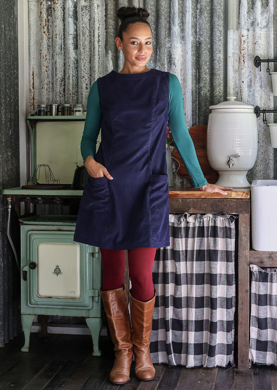 Woman in a navy blue dress standing in a rustic kitchen with checkered curtains and vintage stove.