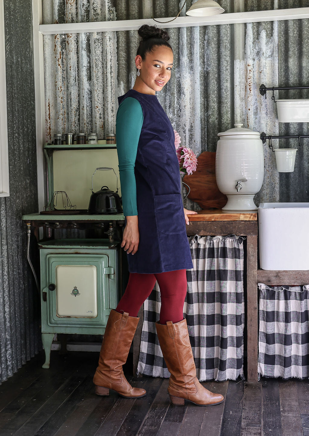 Woman standing in a rustic kitchen with checkered curtains and wooden furniture.
