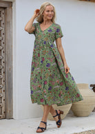 Woman wears 100% cotton button through relaxed fit dress with short sleeves and V-neckline in folk art style floral print on a moss green base.