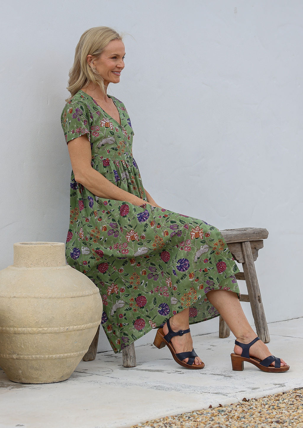 Woman sitting on a wooden bench wearing a lightweight cotton moss green based floral print dress with short sleeves, v-neckline and pockets.