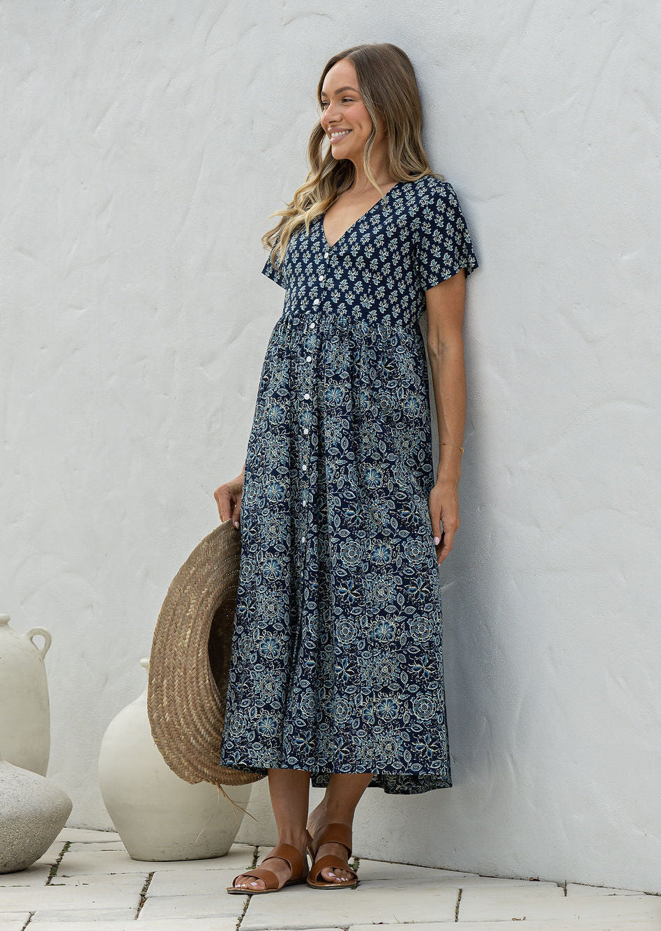 Woman wearing a blue floral dress standing against a white wall with decorative vases.