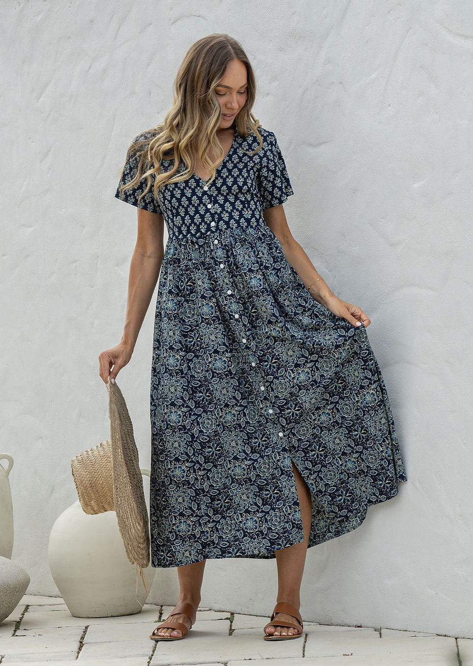 Woman standing in a 100% cotton floral print dress with a V-neckline, short sleeves, pockets, and buttons through to the knee.