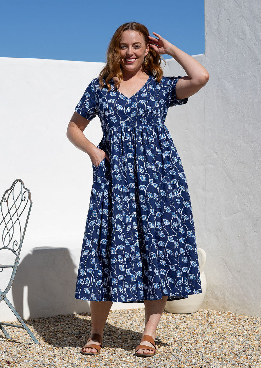 Woman standing in a 100% cotton floral print dress with a V-neckline, short sleeves, pockets, and buttons through to the knee.