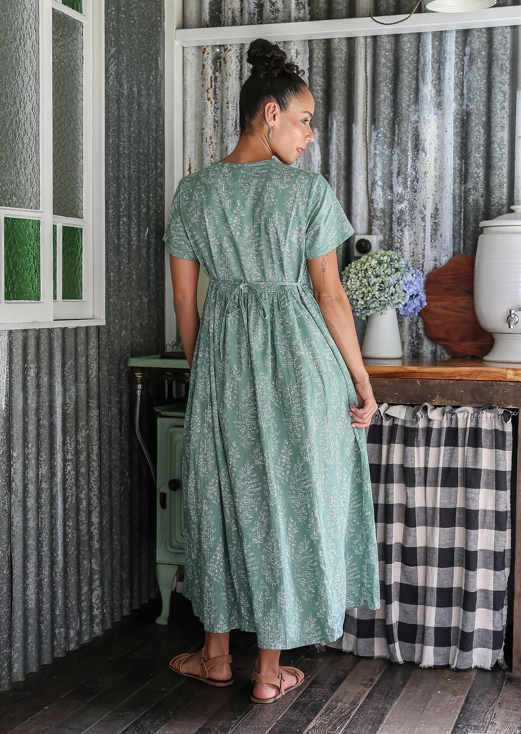 Woman stands backwards wearing a 100% cotton dress with short sleeves, pockets, and a drawstring tie at the back.