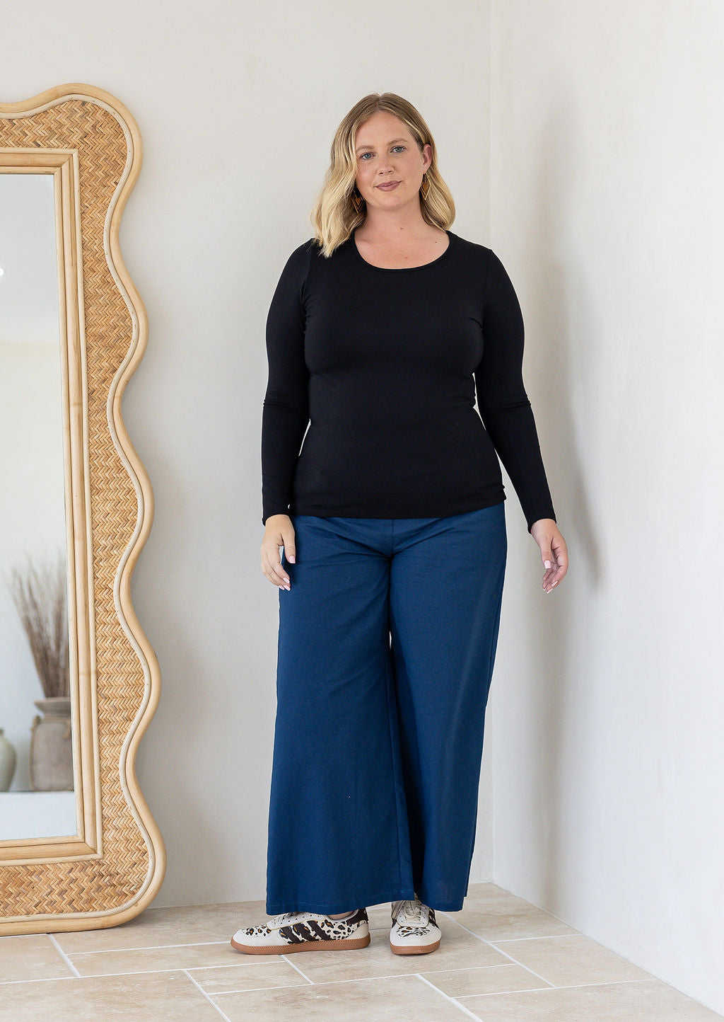A woman modelling a black long-sleeve top and wide-leg cotton blue pants, standing in a bright room with a large wavy-framed mirror on the left.
