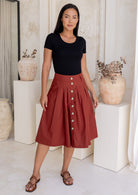 Woman wears rust coloured cotton A-line button through skirt with box pleats and belt loops, paired with a black t-shirt and sandals