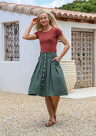 Woman stands with one hand in pockets wearing a tailored knee length cotton skirt with box pleats and belt loops