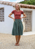 Woman wearing a terracotta basic top and green cotton skirt with button through front and box pleats, standing in front of a white wall with a textured door