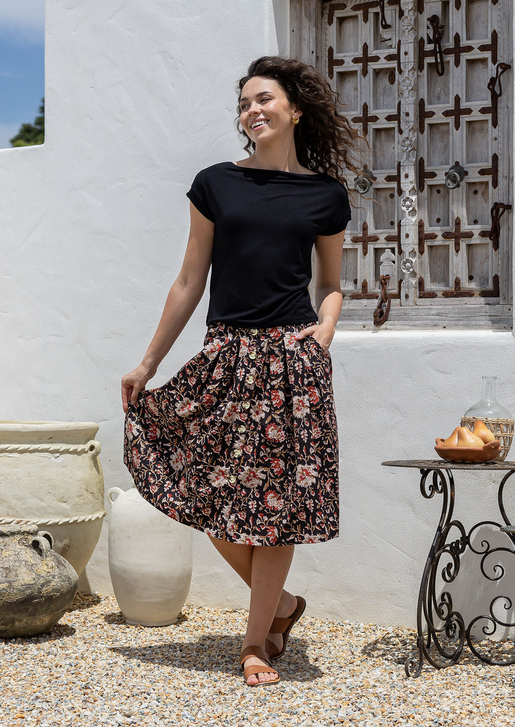 Woman wearing a black top and black based floral print A-line, button through cotton skirt with box pleats, belt loops and pockets