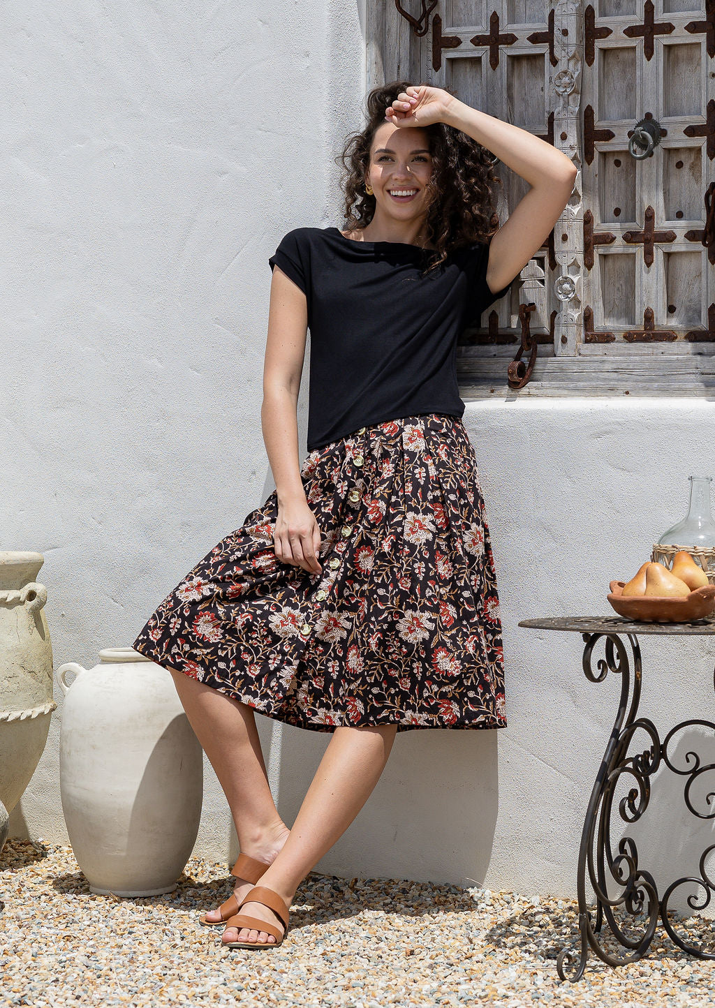 Woman leans on wall in a A line, button through 100% cotton skirt with belt loops, box pleats and elastic back waist.