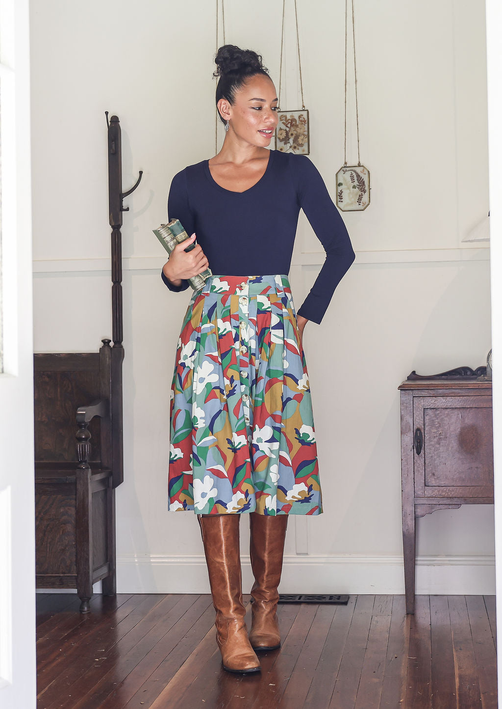 Woman stands wearing a basic top and colourful floral print, button through cotton skirt with box pleats and belt loops