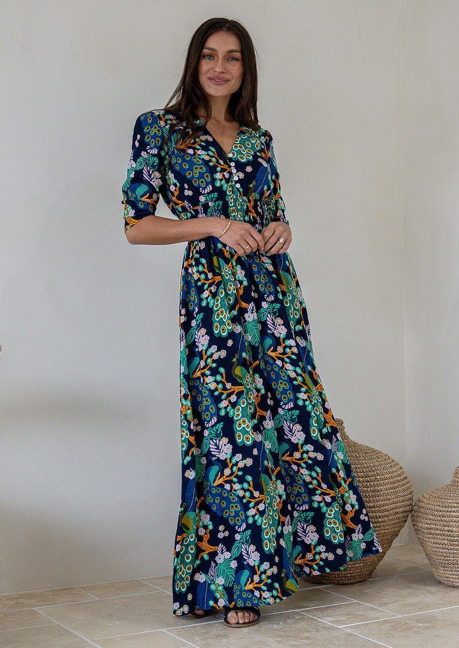 A woman modelling a navy blue rayon maxi dress with 3/4 sleeves, buttoned bodice and shirred waistband, in a colourful floral and peacock print