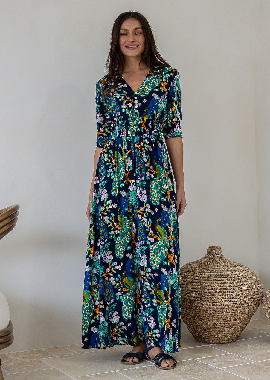 A woman modelling a 3/4 sleeved rayon maxi dress with a vibrant floral and peacock print in shades of navy blue, teal, green, and orange, standing in a minimalist room with a beige wall and woven decorative baskets.