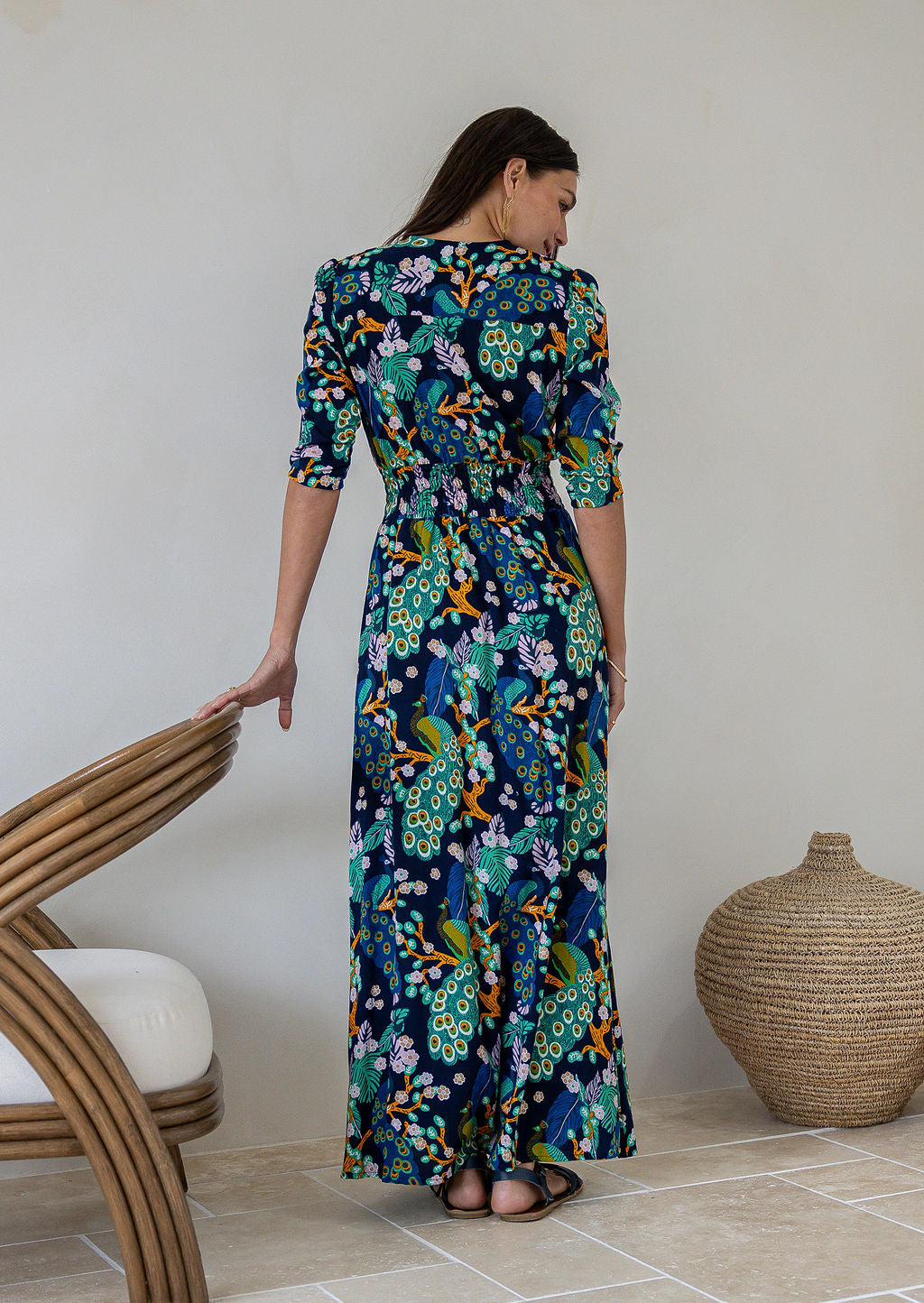 Back view of woman wearing a 3/4 sleeved rayon maxi dress with a vibrant floral and peacock print in shades of teal, green, blue, orange, and purple on a dark navy base.