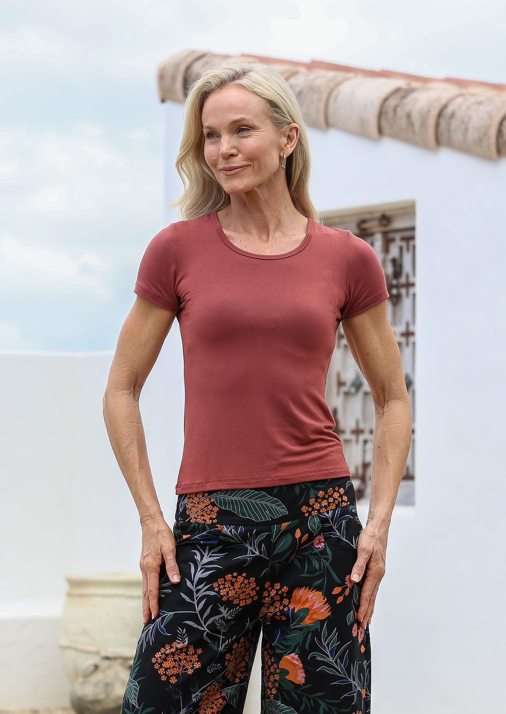 Woman wearing a terracotta fitted t-shirt and floral pants standing against a white wall